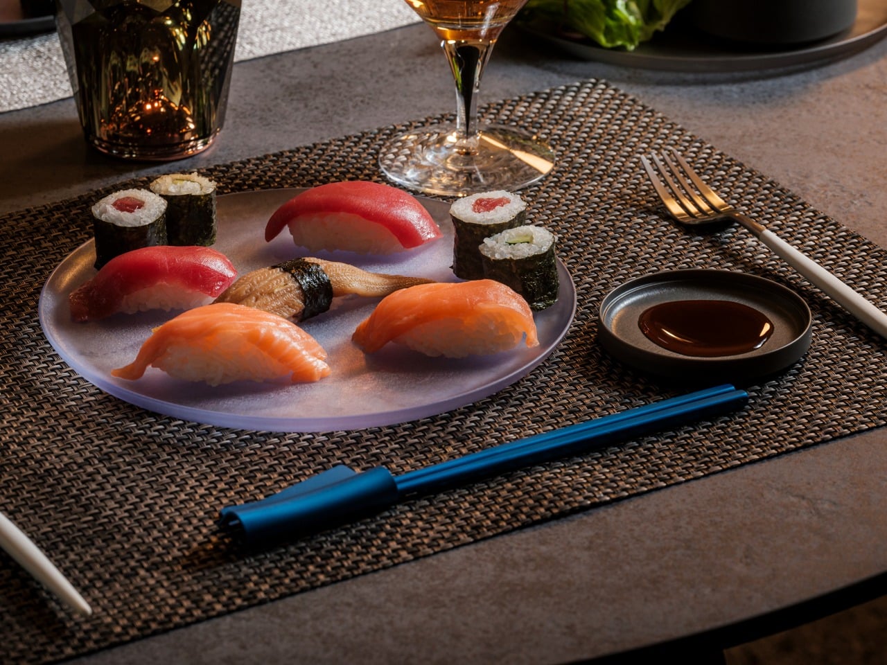 Plate of assorted nigiri and maki sushi on a gray platter, with soy sauce, a wine glass, and blue chopsticks on a woven placemat.