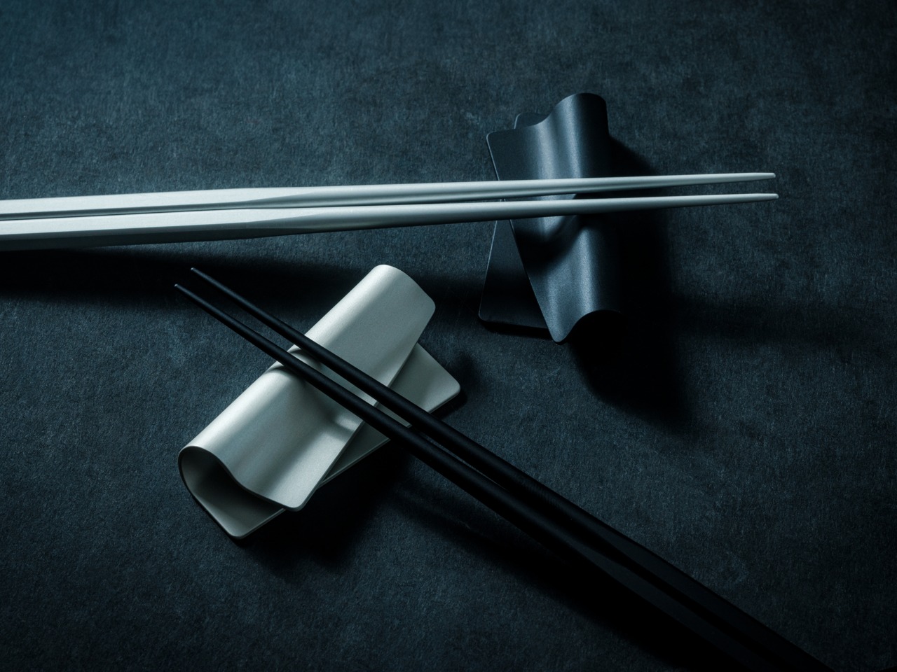 White chopsticks laid across a curved ceramic rest on a dark textured surface with black chopsticks nearby.