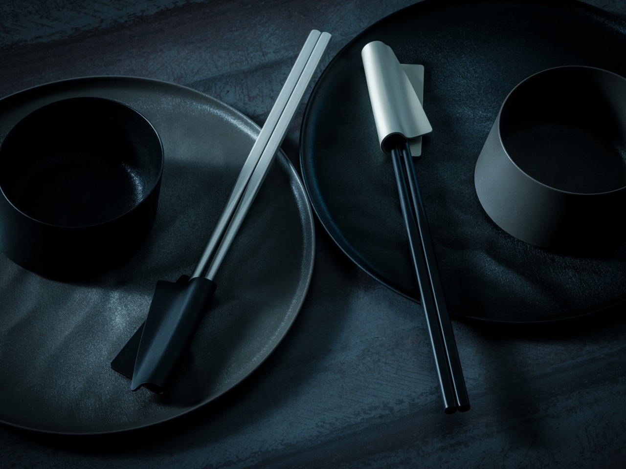 Minimal black tableware setting: two plates with a small black bowl and a pair of chopsticks resting on the plates on a dark textured surface.