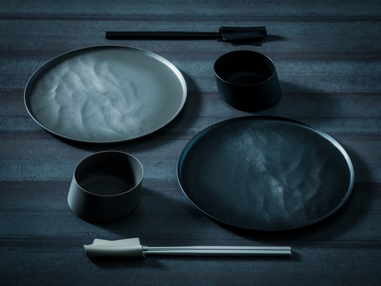 Flat lay of black minimalist dinnerware: two large round plates, two cups, and a pair of chopsticks on a dark textured surface at top and bottom center.