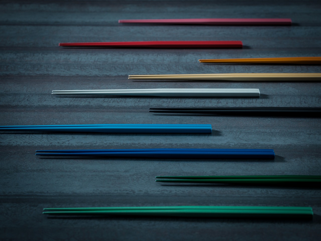 Colorful chopsticks laid diagonally across a dark textured surface, forming a rainbow arrangement.