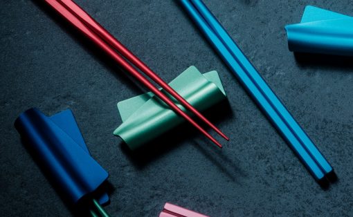 Colorful stationery on a dark textured surface: red chopsticks resting on a green rolled paper with blue strips and pink pieces nearby.