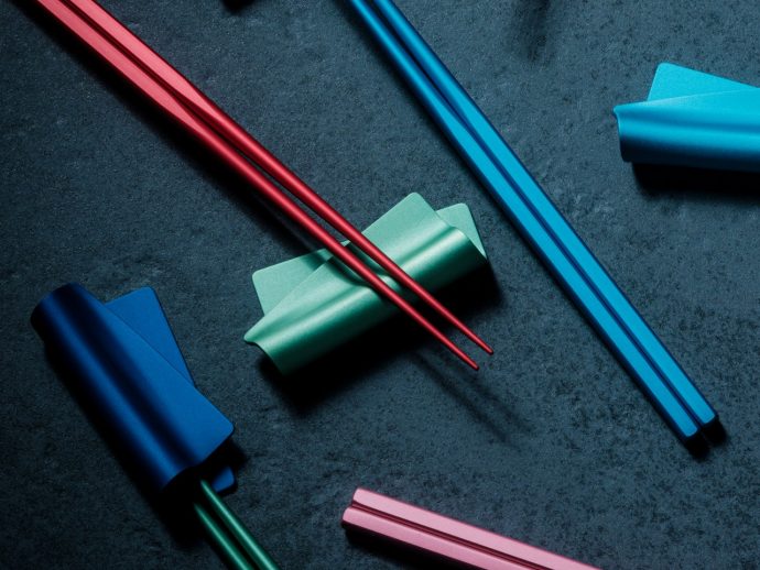 Colorful stationery on a dark textured surface: red chopsticks resting on a green rolled paper with blue strips and pink pieces nearby.