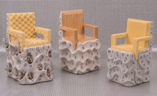 Three decorative ceramic chairs on sculpted bases with calendar-style numbers and blue numerals; left chair has plants, middle is wooden, right is yellow.