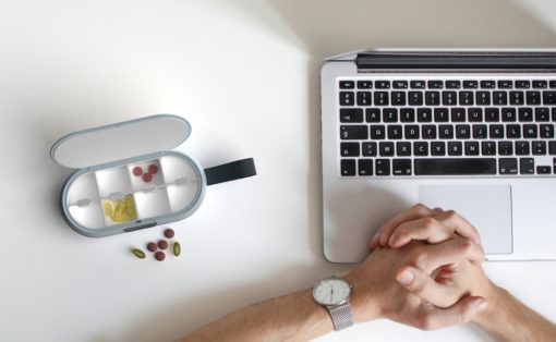 Overhead view of a person clasping hands next to a laptop and a pill organizer on a white desk.