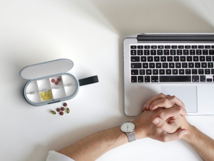 Overhead view of a person clasping hands next to a laptop and a pill organizer on a white desk.