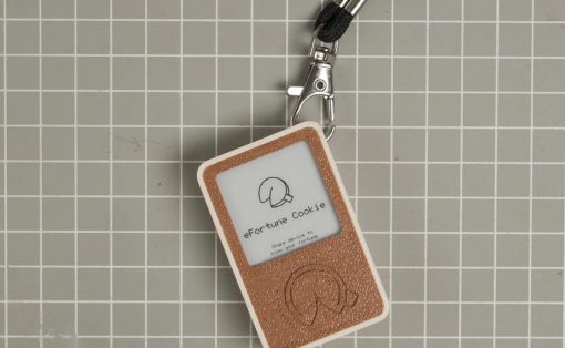 Close-up of a brown textured keychain device labeled 'eFortune Cookie' with a cookie graphic, attached to a black cord on a gray grid background.