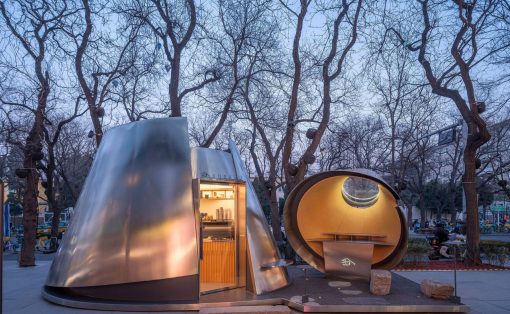 Futuristic metal dome and circular pod sculpture in a tree-filled city park at dusk, with warm interior lighting visible through the doorway and window.