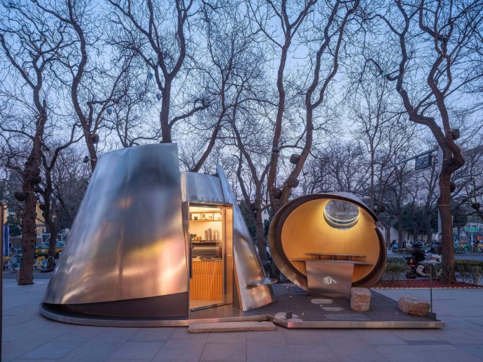 Futuristic metal dome and circular pod sculpture in a tree-filled city park at dusk, with warm interior lighting visible through the doorway and window.
