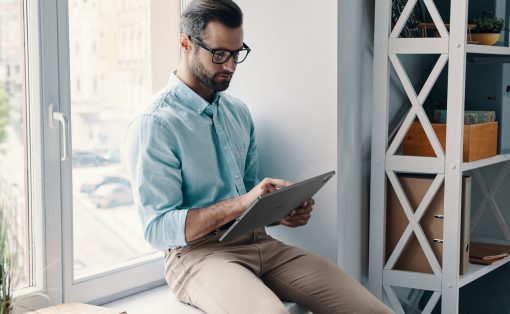 Man with glasses in a light blue shirt sits on a windowsill and uses a tablet in a bright office corner.