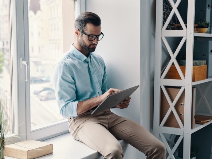 Man with glasses in a light blue shirt sits on a windowsill and uses a tablet in a bright office corner.