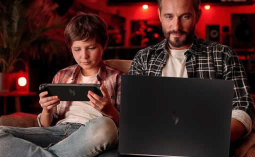 Father and son relax on a couch, the boy playing a handheld game while the man uses a laptop nearby in a red-lit room.