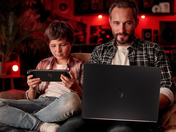 Father and son relax on a couch, the boy playing a handheld game while the man uses a laptop nearby in a red-lit room.