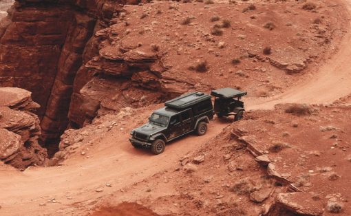 Black four‑wheel‑drive SUV with a trailer navigating a dusty red canyon road on a rugged trail.