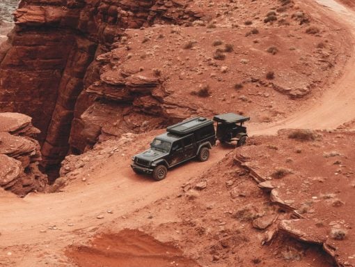 Black four‑wheel‑drive SUV with a trailer navigating a dusty red canyon road on a rugged trail.