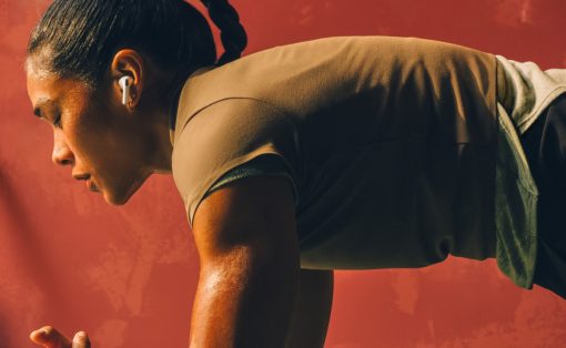 Woman in a high plank position with forearms on dumbbells, wearing wireless earbuds against a red wall.