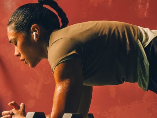Woman in a high plank position with forearms on dumbbells, wearing wireless earbuds against a red wall.