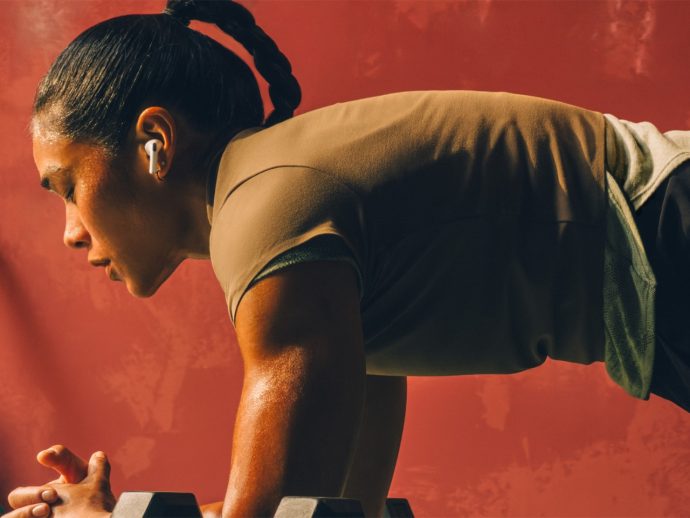 Woman in a high plank position with forearms on dumbbells, wearing wireless earbuds against a red wall.