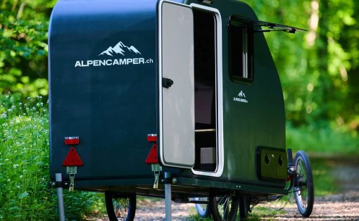 Dark green Alpencamper teardrop camper with its door open, parked on a forest trail with stabilizing legs.