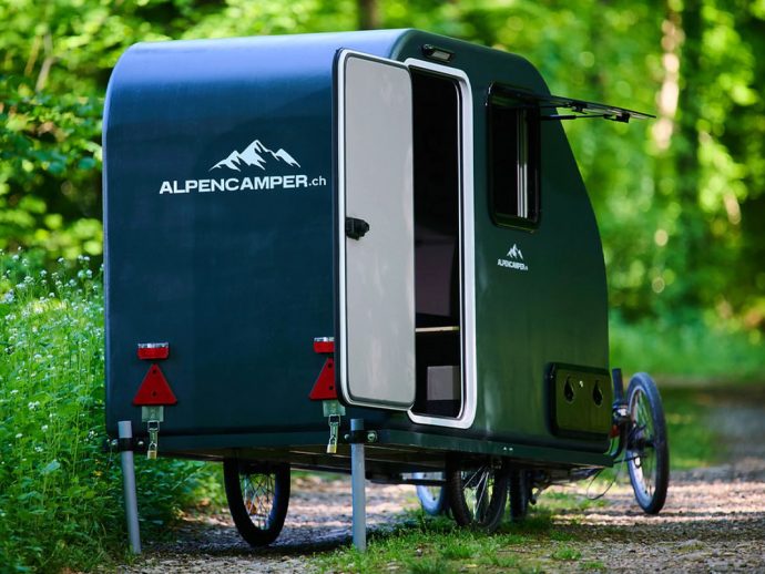 Dark green Alpencamper teardrop camper with its door open, parked on a forest trail with stabilizing legs.
