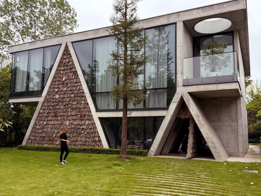 Modern angular concrete house with large glass windows, shingled triangular wall, and a person walking on a green lawn beside a tree.