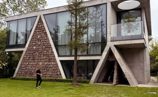 Modern angular concrete house with large glass windows, shingled triangular wall, and a person walking on a green lawn beside a tree.