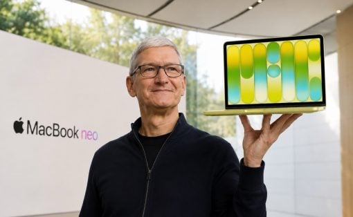 Smiling man with glasses holds up a bright gradient laptop in a showroom, MacBook Neo signage visible in the background.