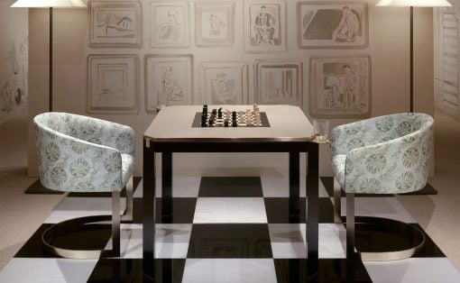 Two patterned armchairs face a square chess table on a black-and-white checkered floor, with lamps and framed wall art behind.