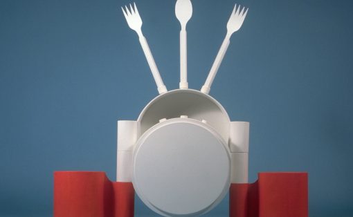Abstract white sculpture resembling a person with a spoon as head and two forks raised like arms, set against a blue backdrop with red cylinders at the base