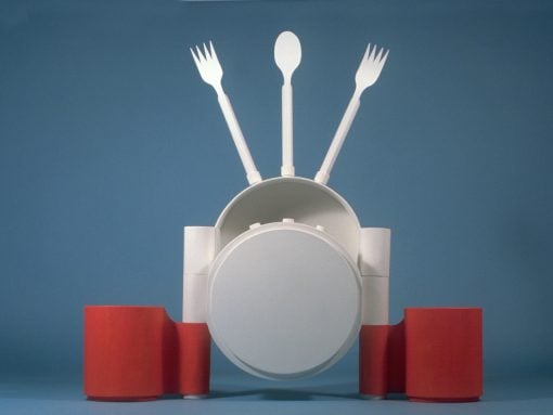 Abstract white sculpture resembling a person with a spoon as head and two forks raised like arms, set against a blue backdrop with red cylinders at the base