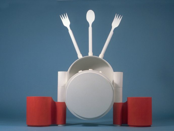 Abstract white sculpture resembling a person with a spoon as head and two forks raised like arms, set against a blue backdrop with red cylinders at the base