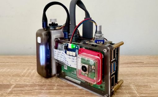 DIY single-board computer in a metal frame with a webcam-style camera module, green power LED, and exposed USB ports on a wooden table.