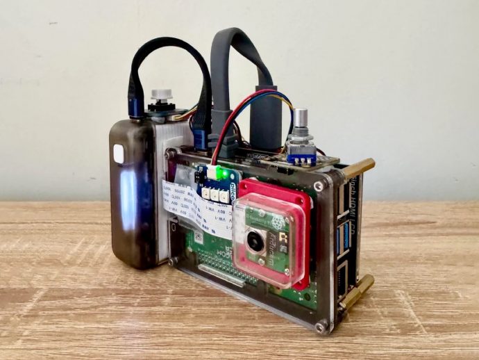 DIY single-board computer in a metal frame with a webcam-style camera module, green power LED, and exposed USB ports on a wooden table.