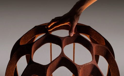 Close-up of a hand lifting a rust-colored, hollow geometric lattice sphere with circular openings.