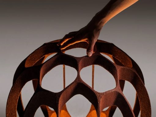 Close-up of a hand lifting a rust-colored, hollow geometric lattice sphere with circular openings.
