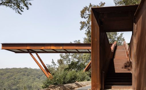 Rusty metal stairway and platform with outdoor hillside view, leading up to a walkway over a forested landscape.