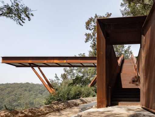 Rusty metal stairway and platform with outdoor hillside view, leading up to a walkway over a forested landscape.