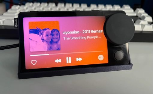 Portable music player on a stand displaying a Spotify screen with a photo of two girls on the left and song title on the right, plus playback controls below. The device sits in front of a keyboard.