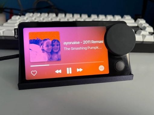 Portable music player on a stand displaying a Spotify screen with a photo of two girls on the left and song title on the right, plus playback controls below. The device sits in front of a keyboard.