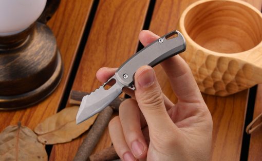 Close-up of a hand holding a small gray folding knife with a partially serrated blade on a wooden table.