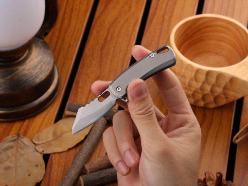 Close-up of a hand holding a small gray folding knife with a partially serrated blade on a wooden table.