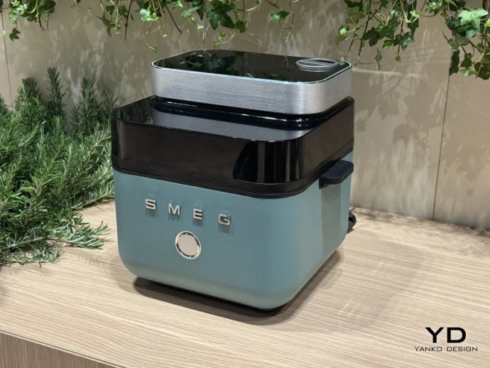 Teal retro-style SMEG appliance with a glossy black top on a light wood counter, greenery in the background.