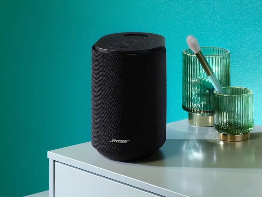 Black cylindrical Bose smart speaker with fabric cover on a white table, beside two green ribbed glasses and a brush against a teal wall.