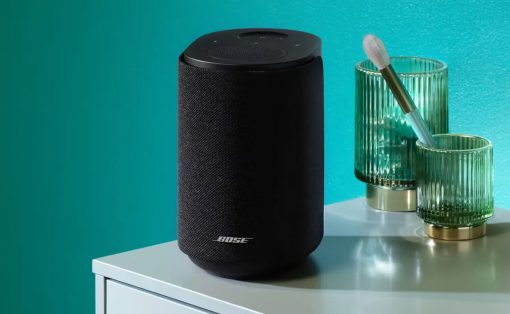 Black cylindrical Bose smart speaker with fabric cover on a white table, beside two green ribbed glasses and a brush against a teal wall.