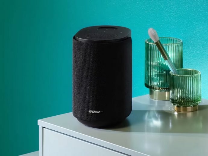 Black cylindrical Bose smart speaker with fabric cover on a white table, beside two green ribbed glasses and a brush against a teal wall.