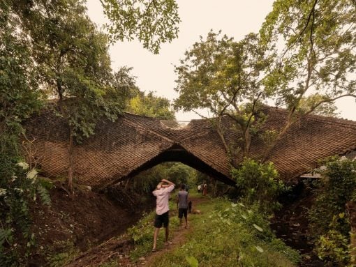 People walk along a muddy path under a large, thatched arch bridge set in a lush, tropical area.