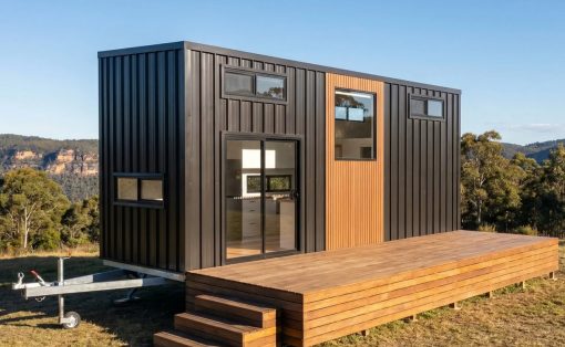 Modern tiny house on a trailer with black metal siding, wooden deck, and steps in a rural landscape.