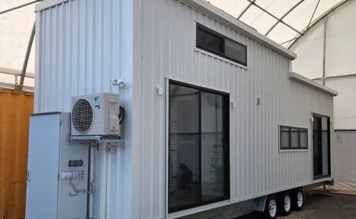 White modular trailer house on a multi-axle trailer with large glass doors and windows, and an outdoor HVAC unit mounted on the side.