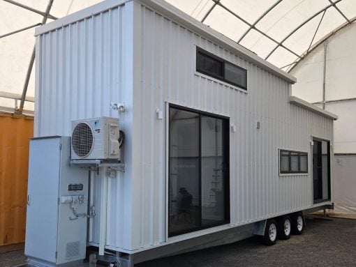 White modular trailer house on a multi-axle trailer with large glass doors and windows, and an outdoor HVAC unit mounted on the side.