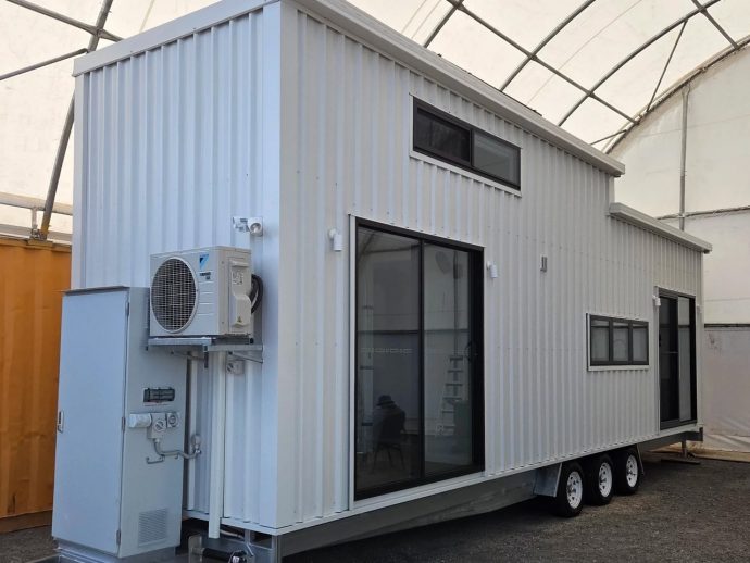 White modular trailer house on a multi-axle trailer with large glass doors and windows, and an outdoor HVAC unit mounted on the side.